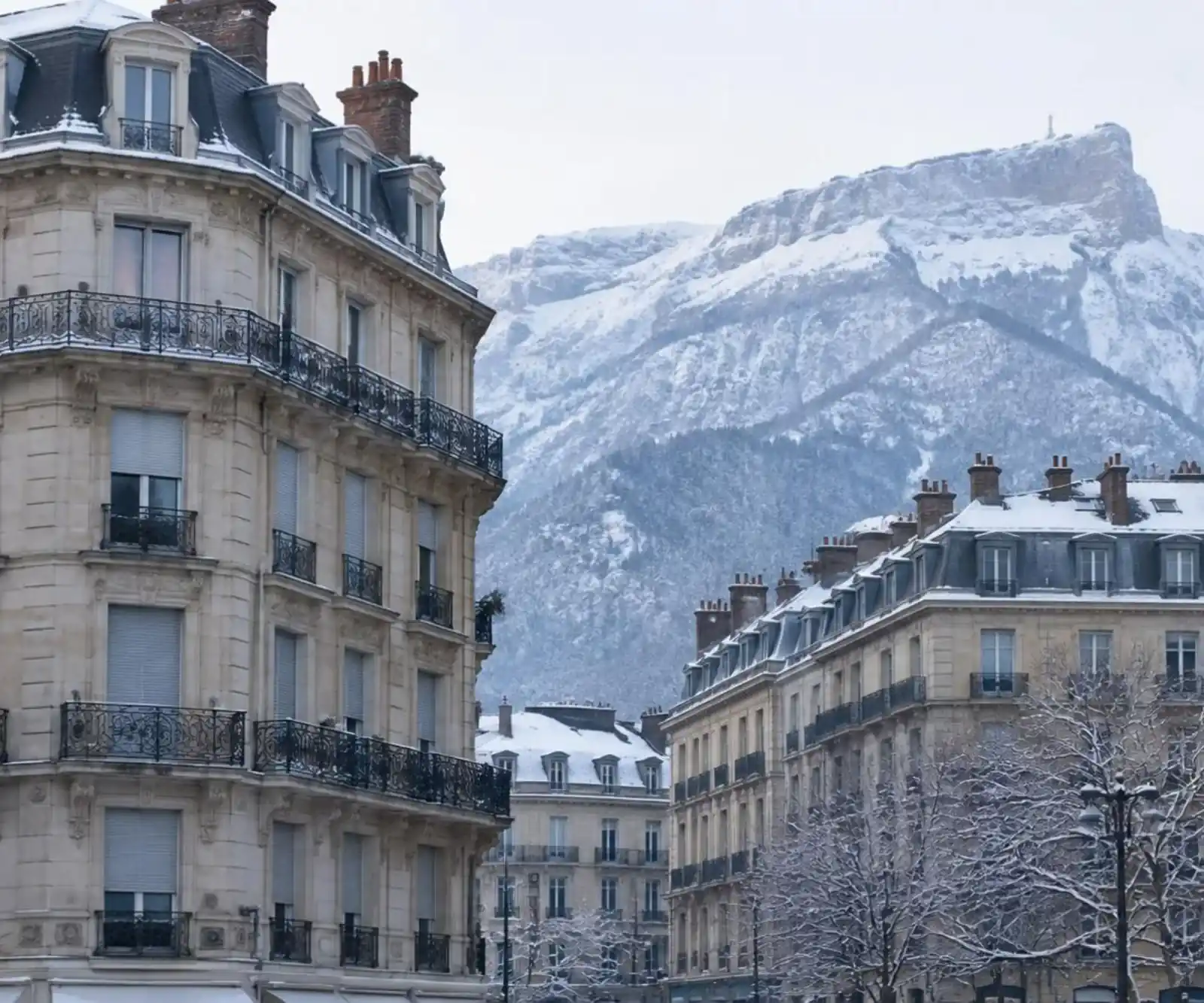 Immeubles grenoblois avec balcons et volets fermés, montagnes enneigées derrière